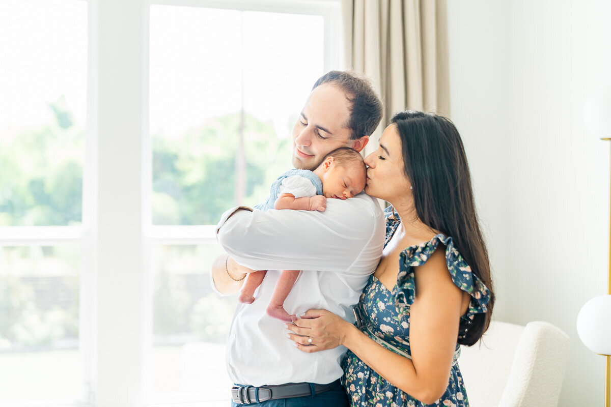 a mother and father stand in front of a window at their Leander home. The father holds their newborn while the mother kisses the infant on the forehead.