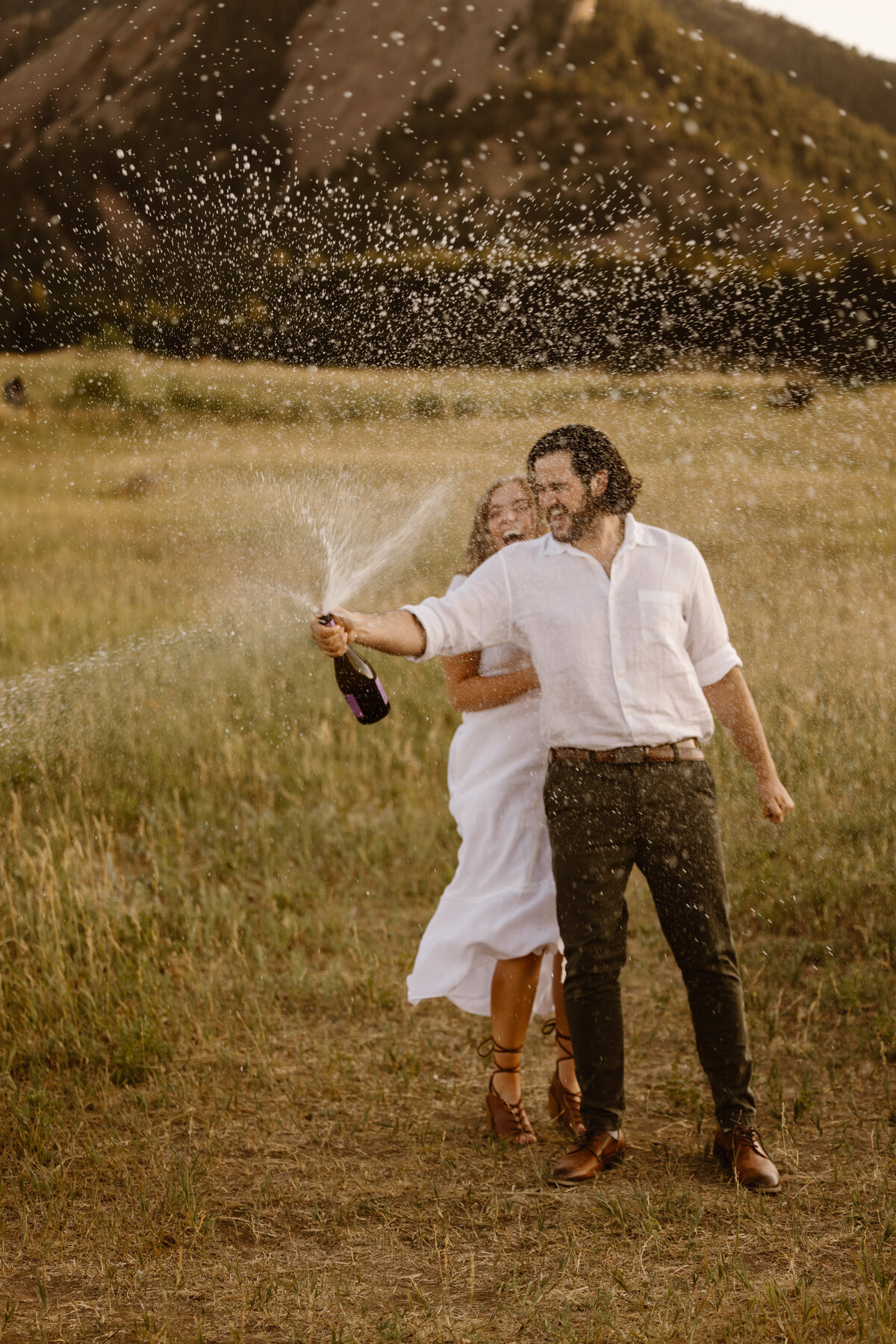 A couple in white outfits laughs joyfully as they pop a champagne bottle in an open field with the Flatirons in the background, surrounded by a spray of bubbles.