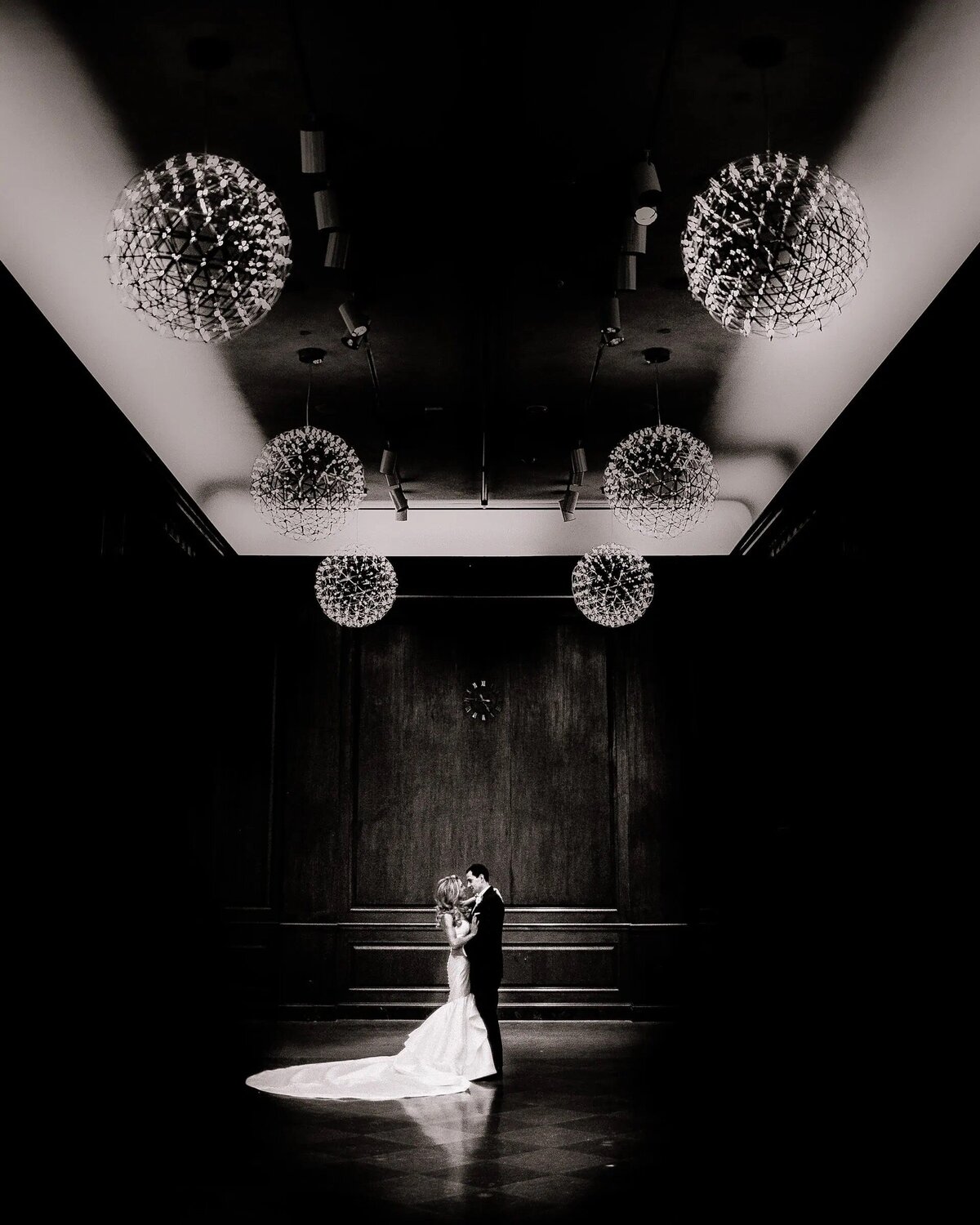 A couple stands embraced in the spotlight, surrounded by darkness and the grandeur of ornate chandeliers above.