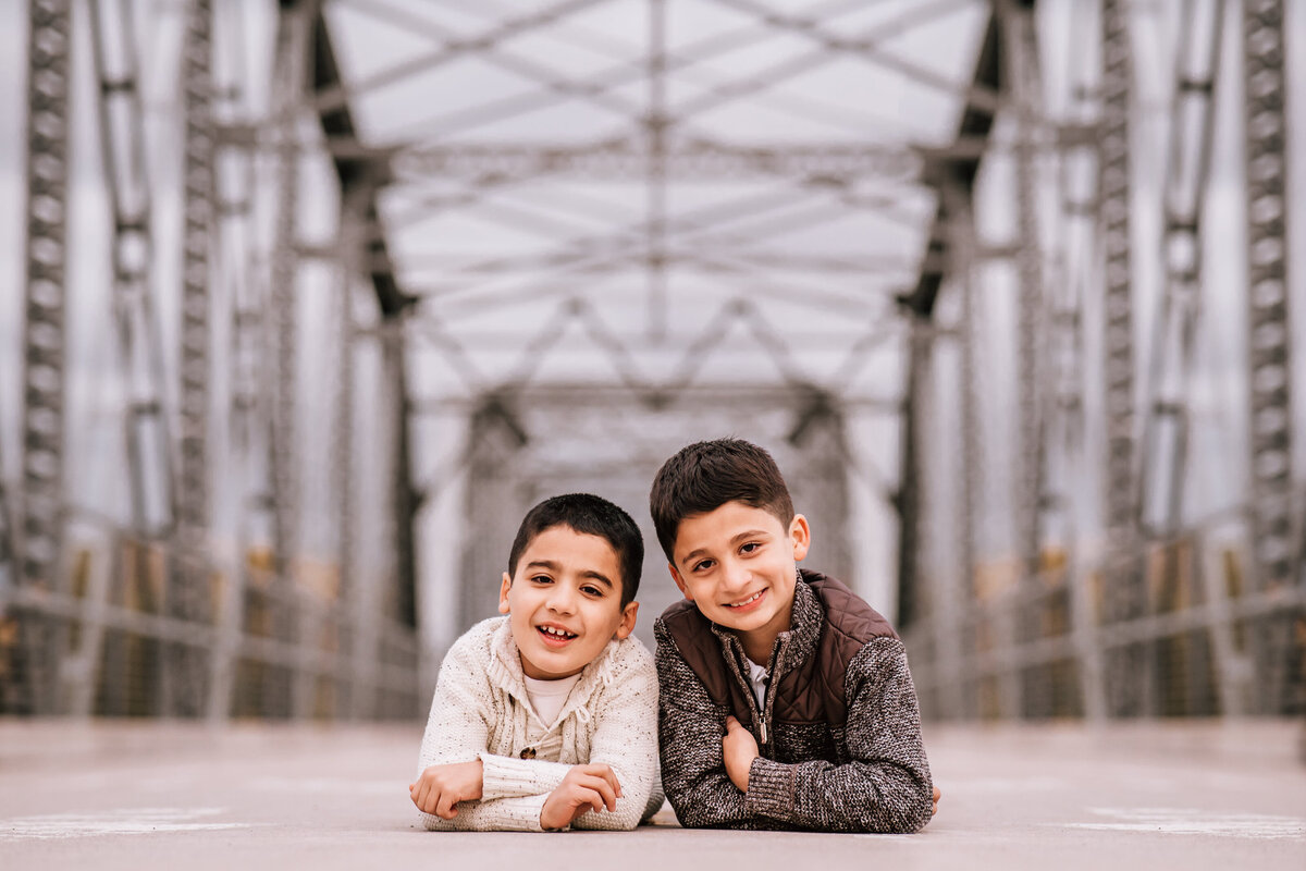 Two young boys lie on their stomachs, smiling at the camera, on a metal bridge with an intricate framework in the background. Both boys are dressed in cozy sweaters, enjoying the moment.