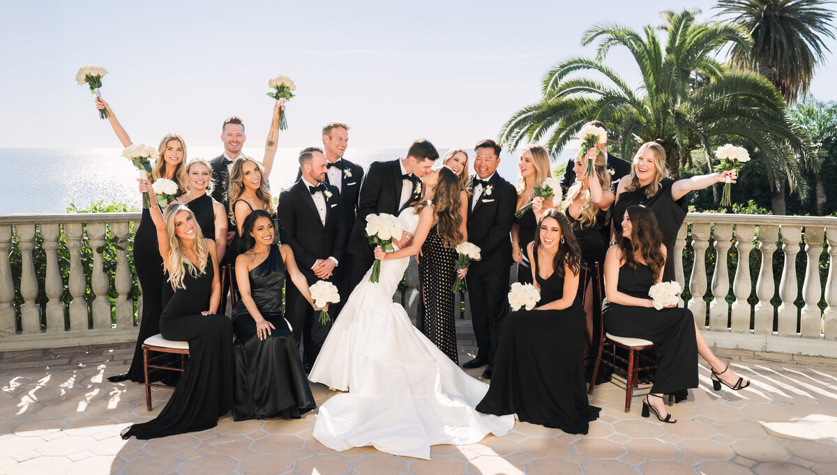 A joyful bridal party celebrating at a luxurious Bel-Air Bay Club wedding in Malibu. The bride and groom share a romantic kiss, surrounded by bridesmaids in black dresses and groomsmen in classic tuxedos, with a stunning ocean view in the background. Captured by a Los Angeles wedding photographer.