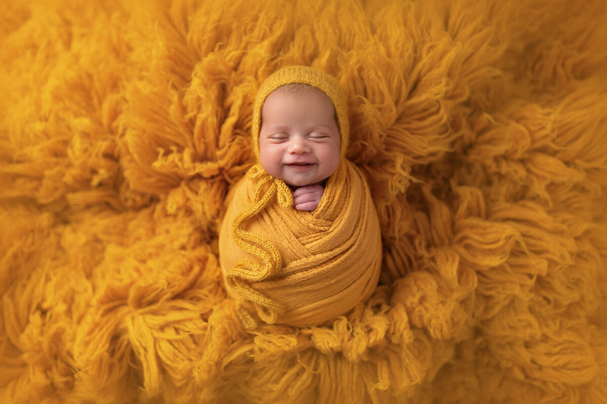 Newborn baby smiling while wrapped in mustard yellow on a fluffy textured golden backdrop.