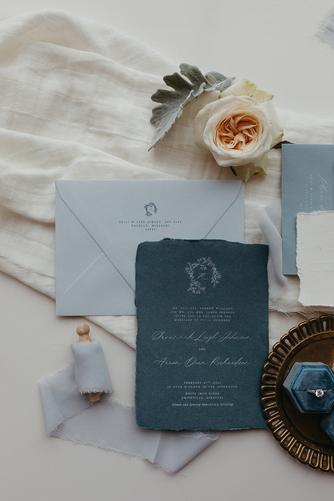 An assortment of blue and gray wedding stationery with white cursive font atop fabric, with a wedding ring in blue velvet box