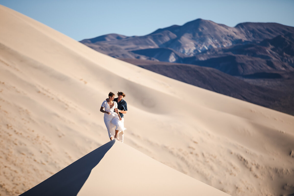 bride and groom embrace standing on tip of sand dune hair blowing in the wind