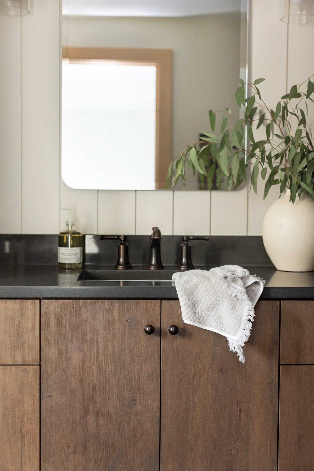 Natural wood bathroom vanity with dark grey countertops and antique bronze widespread faucet. Grey and white towel draped over the sink. Bottle of soap to the left of the sink. Large beige vase with green florals to the right of the sink. Off-white shiplap on the wall behind the vanity with a mirror.