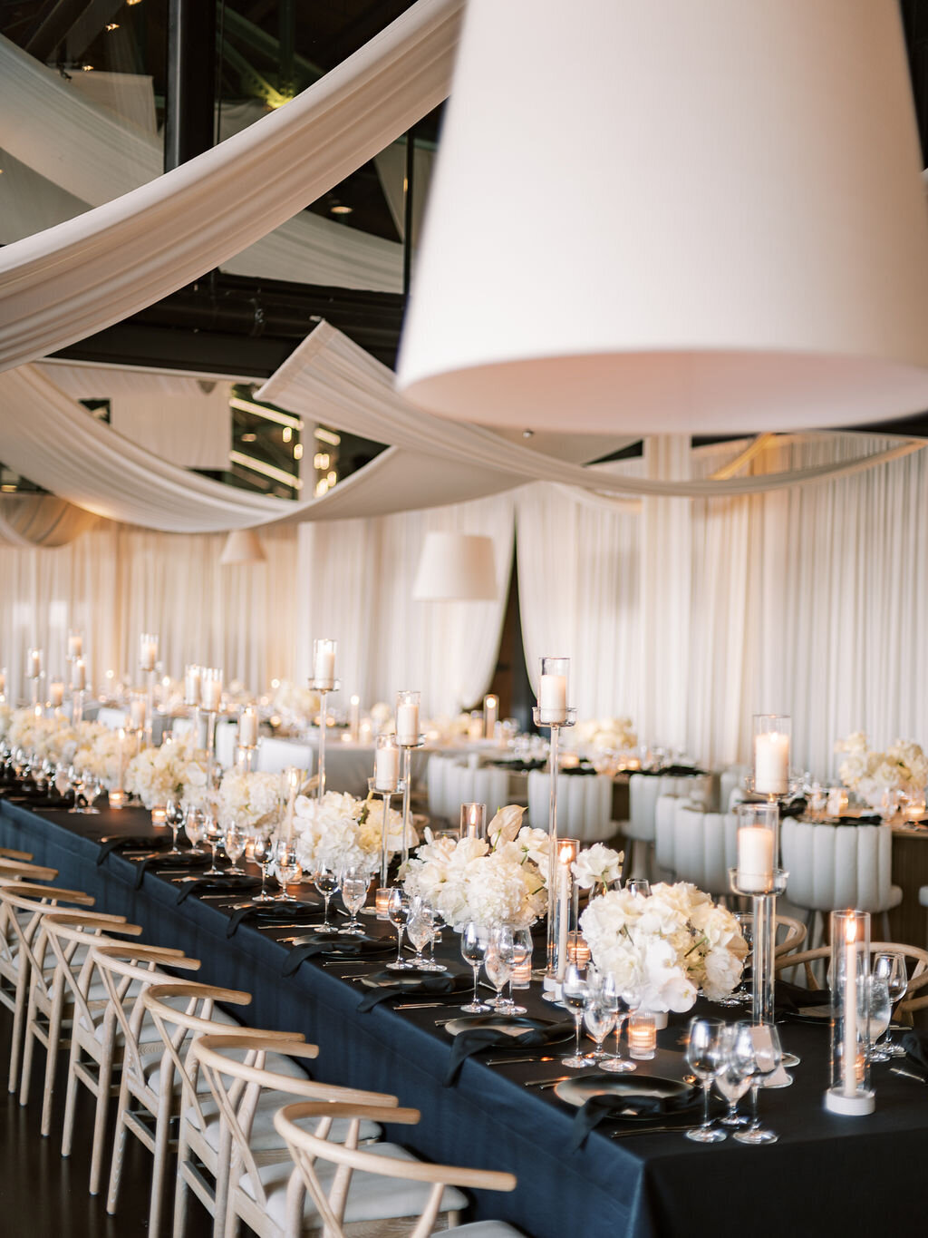 A long wedding reception table adorned with white floral centerpieces, candlelight, and draped fabric overhead.