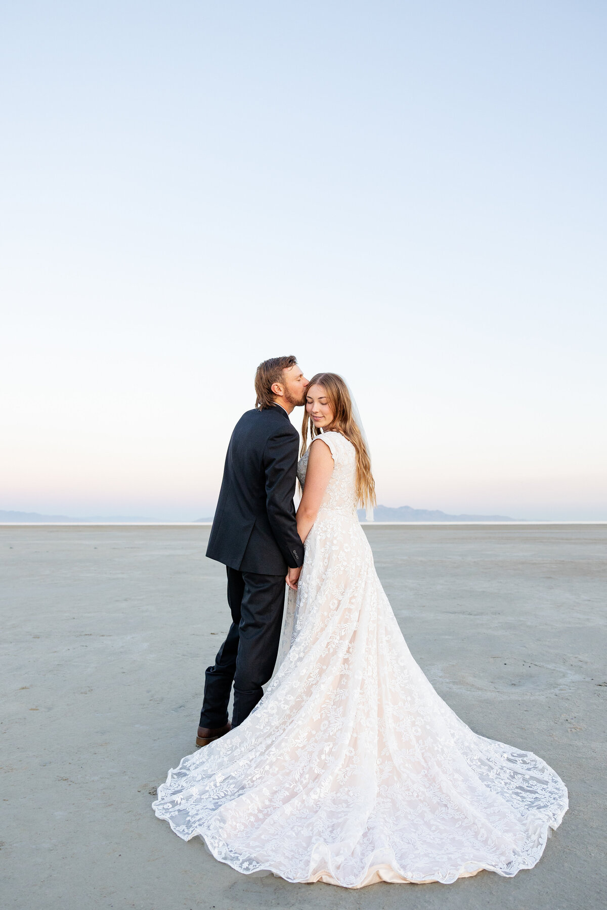 Groom in a black suit kisses the bride’s forehead as she smiles softly, with a vast landscape behind them.