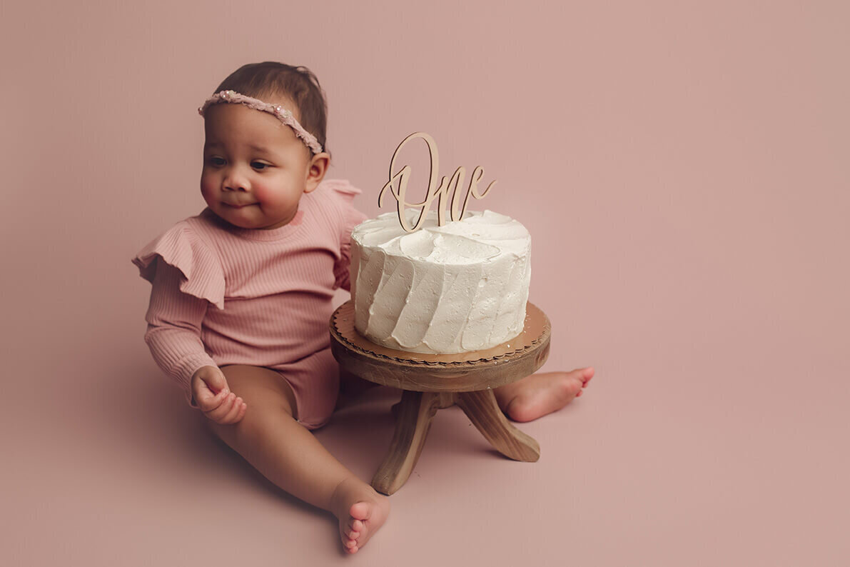 a one year old girl looking away from her white cake with a pick that says one