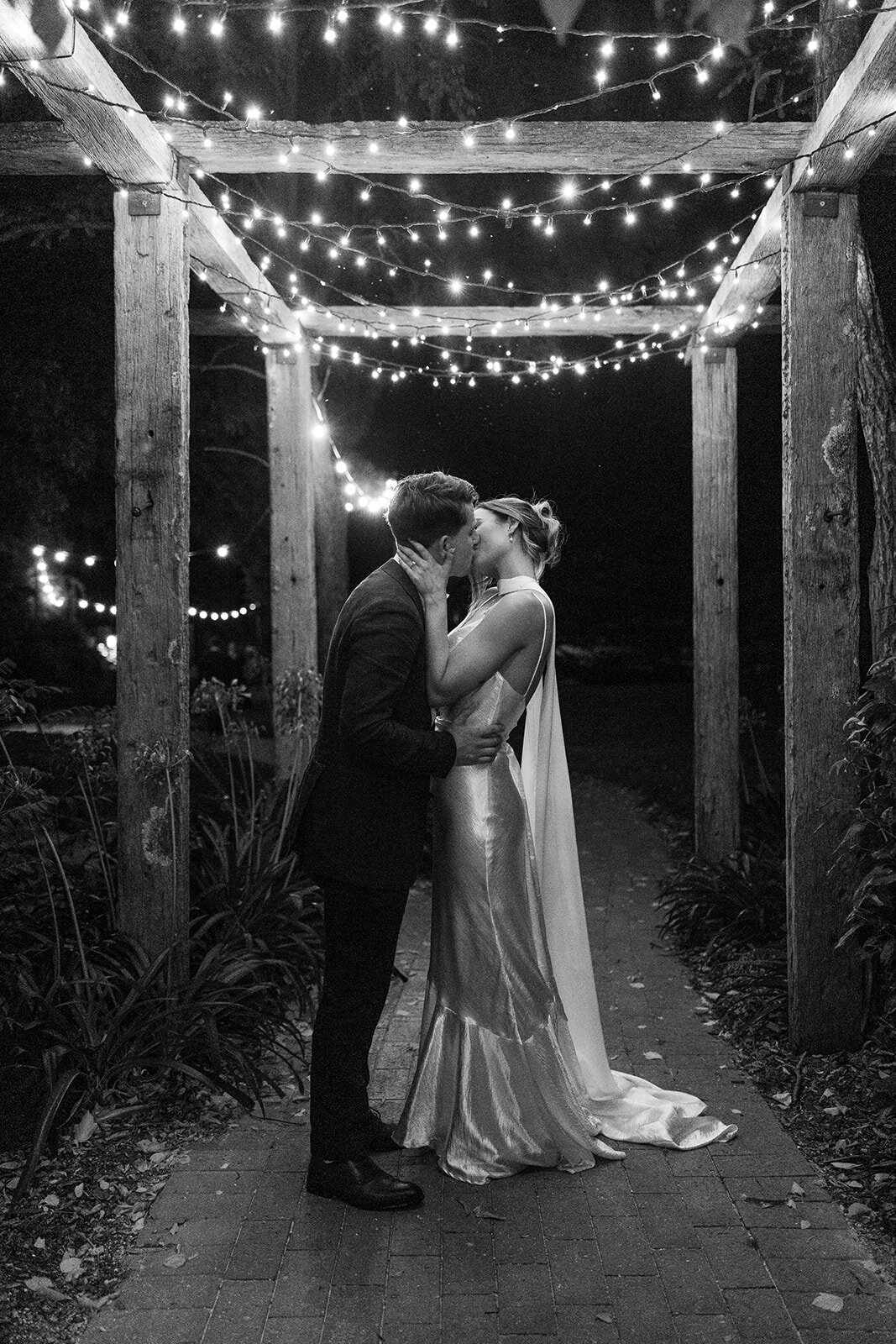 A black and white image of bride and groom embracing under fairy lights on their wedding night. Taken at the entrance of Poachers Pantry.