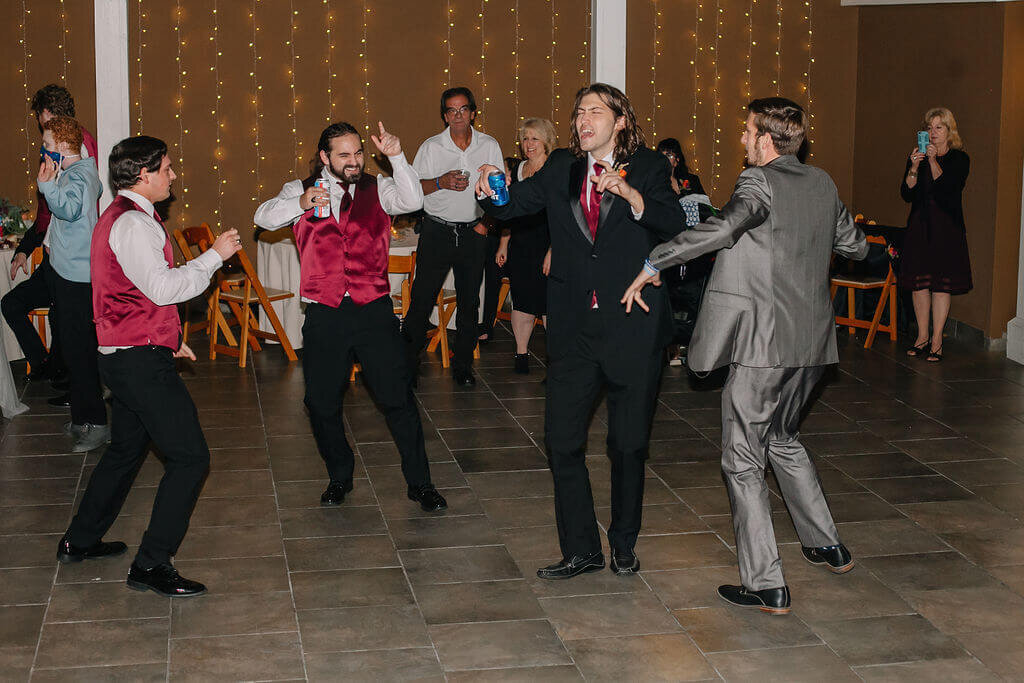 A group of men in suits and red vests dance enthusiastically on a tiled floor at a party, while guests in the background watch and hold drinks. String lights hang on the wall behind them.
