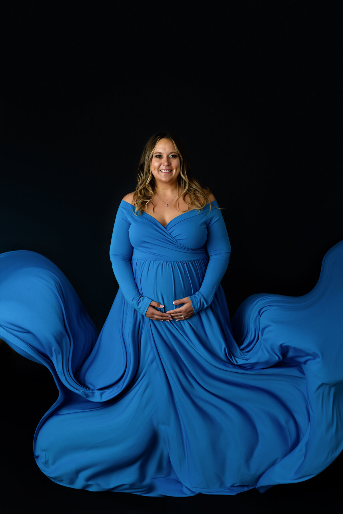 A happy mom to be in a blue maternity gown stands in a studio as her train flows all around her in the wind