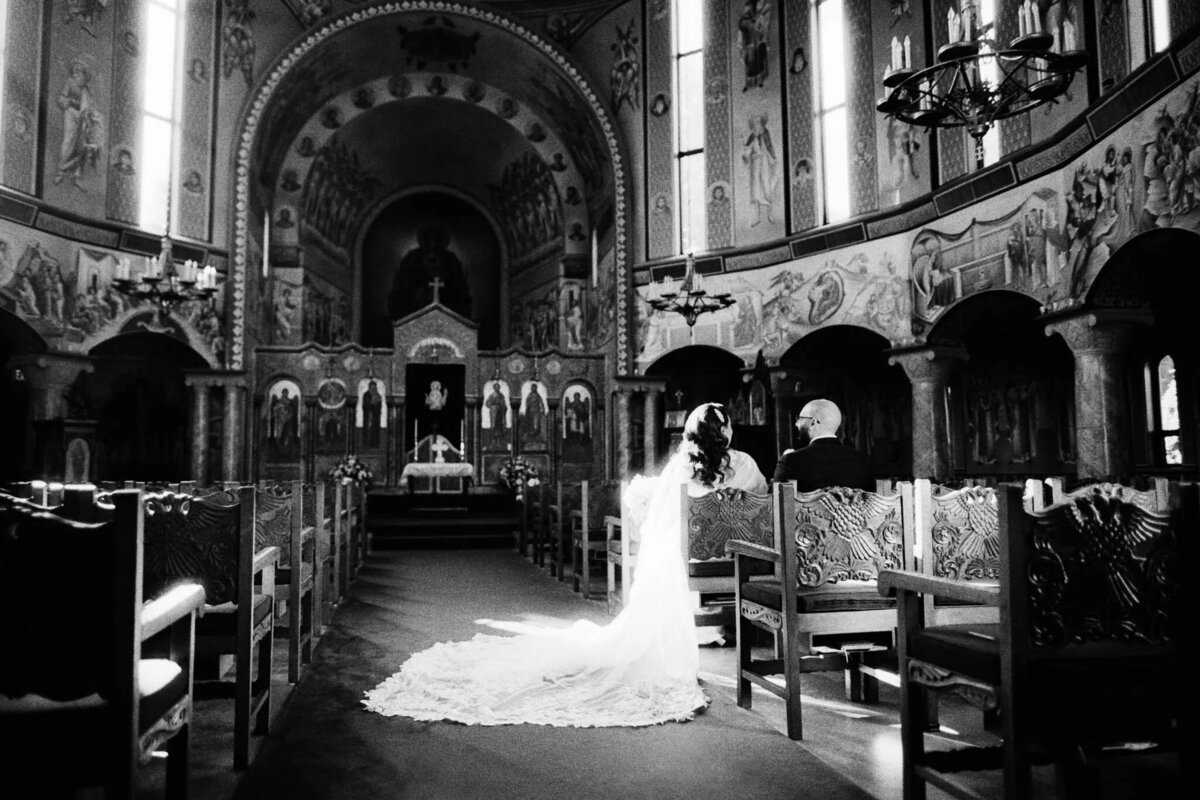 A bride and groom sit together in ornate wooden pews inside a grand, elaborately decorated church with high arched ceilings and detailed murals—sunlight illuminating the bride’s long train, perfectly captured by an experienced NJ wedding photographer.