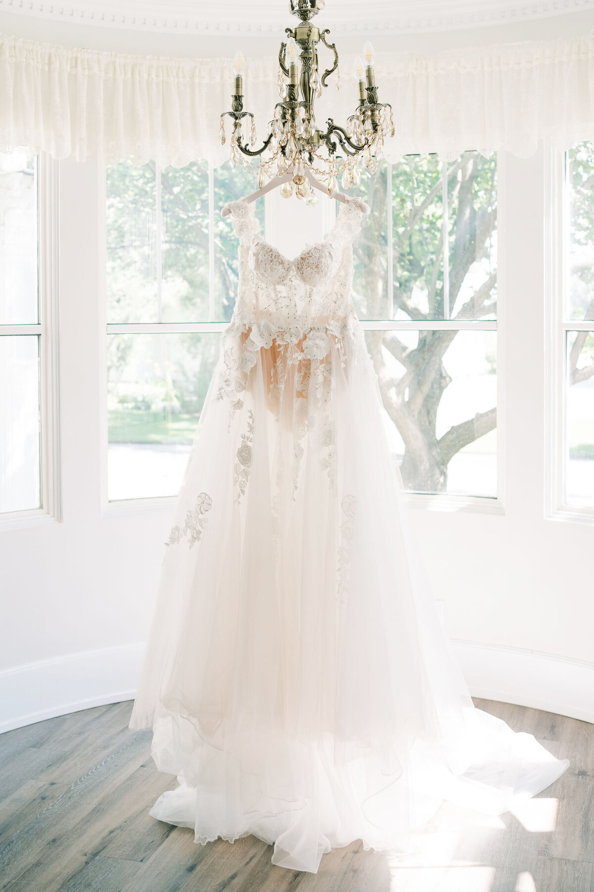 A lace and tulle wedding dress hangs from a chandelier in front of large windows with sheer curtains, illuminated by natural light, showcasing the impeccable wedding design for the upcoming wedding in Canada.