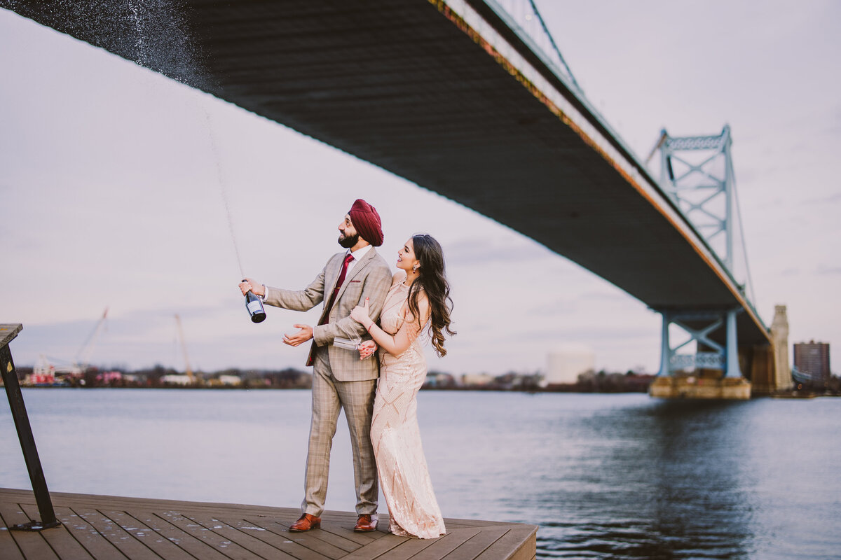 Engaged couple spraying a bottle of champagne in the air at race street pier with the bridge in the background in philadelphia