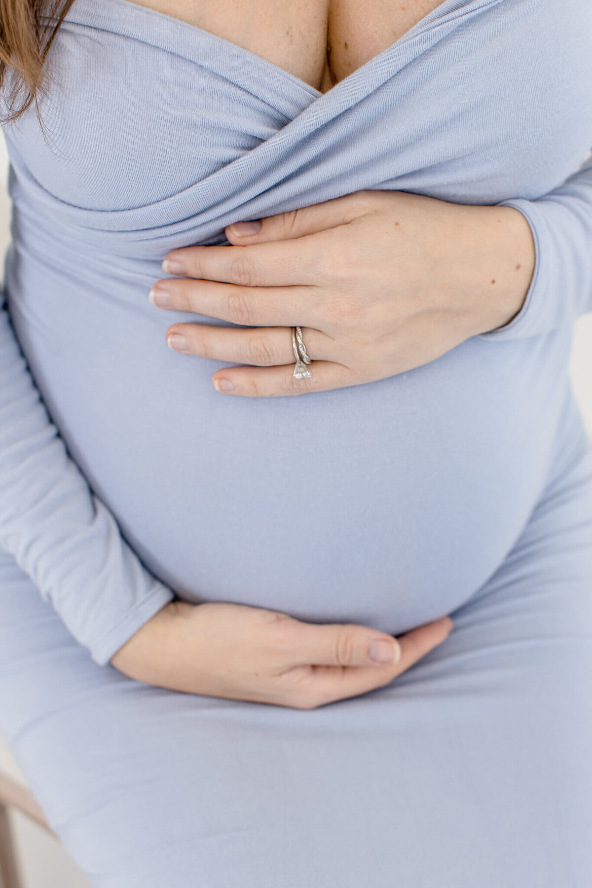A woman in a light blue dress holds her pregnant belly with her hands, one adorned with a ring. The dress has long sleeves and a wrap-style bodice. The focus is on the belly and hands, conveying a sense of anticipation and care.