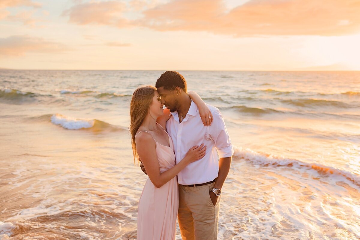 Woman in pastel pink dress wraps her arms around her husband, the newly engaged couple smiles at the beach as they are photographed by Love + Water on Maui