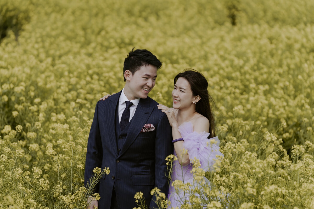 the bride back hugging the groom while smiling at each other in the yellow canola fields in jeju island