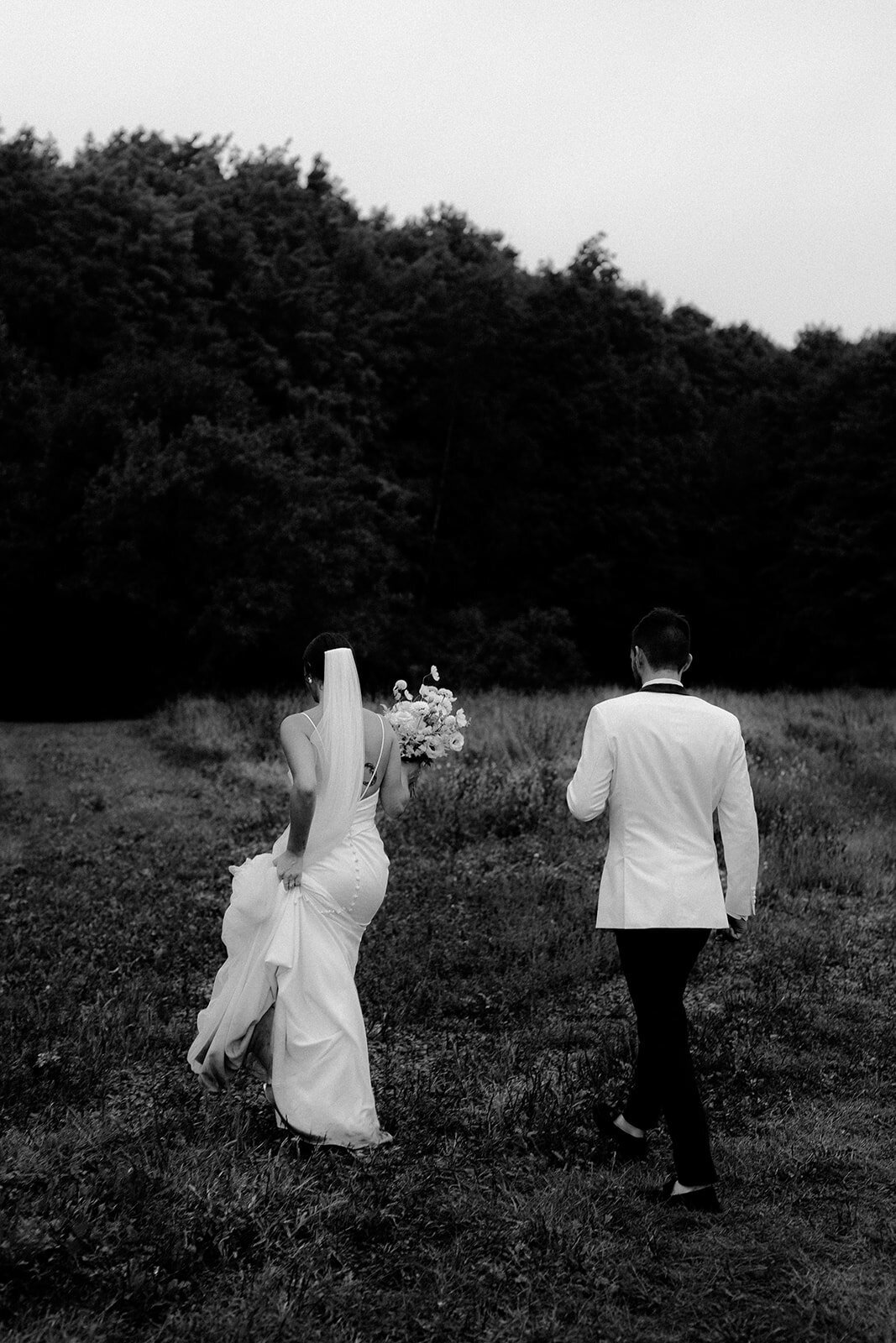 A bride and groom walk through a field at a Montreal wedding, holding a large flower bouquet.