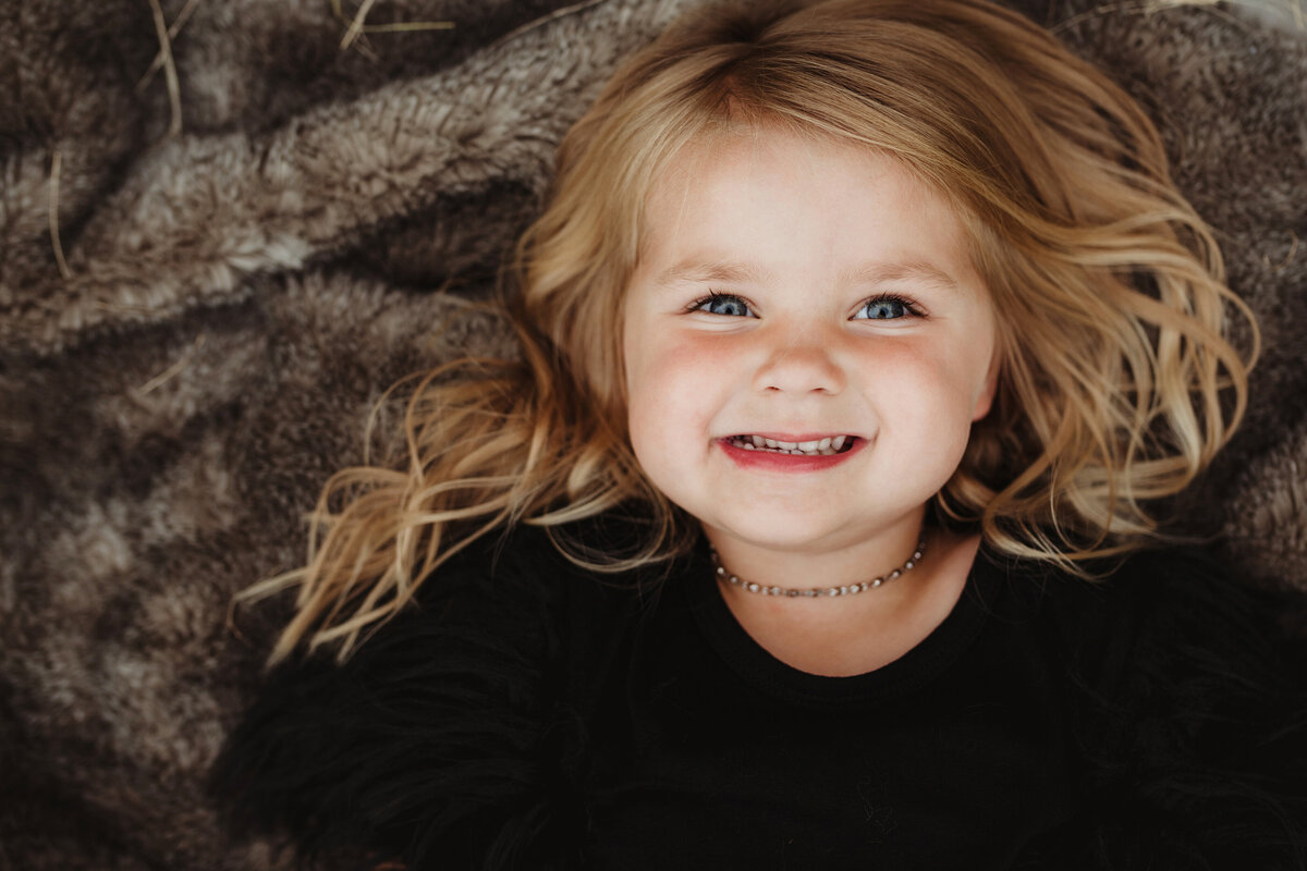 A little girl is lying on a blanket, looking up and smiling at the camera.