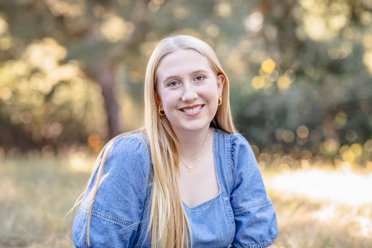 A close-up of a young blonde woman wearing a denim blue shirt smiling at the camera while sitting in the grass at a park.