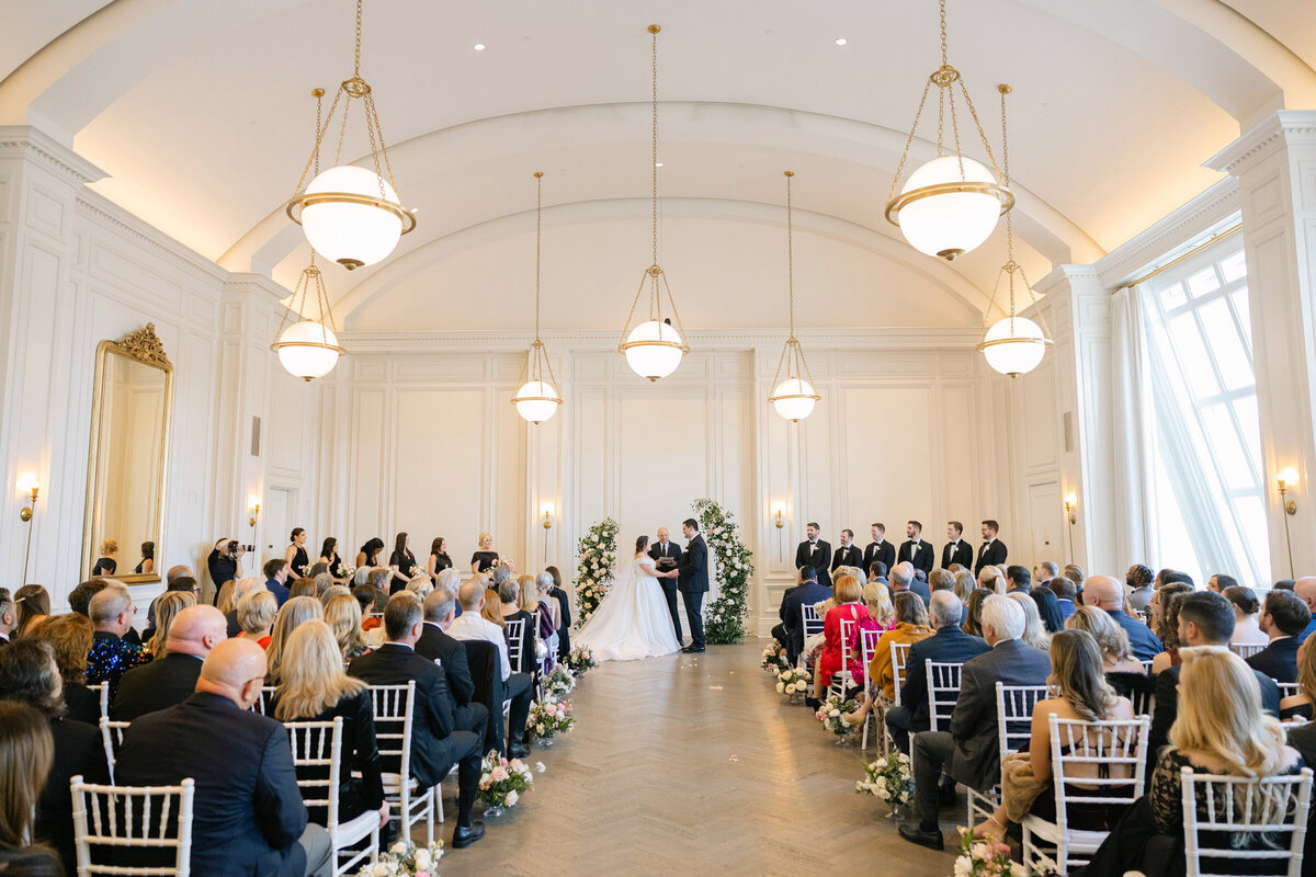 Courtney and Jack holding hands during their wedding ceremony at The Adolphus in Dallas, framed by a floral arch that highlights the elegant venue setting.