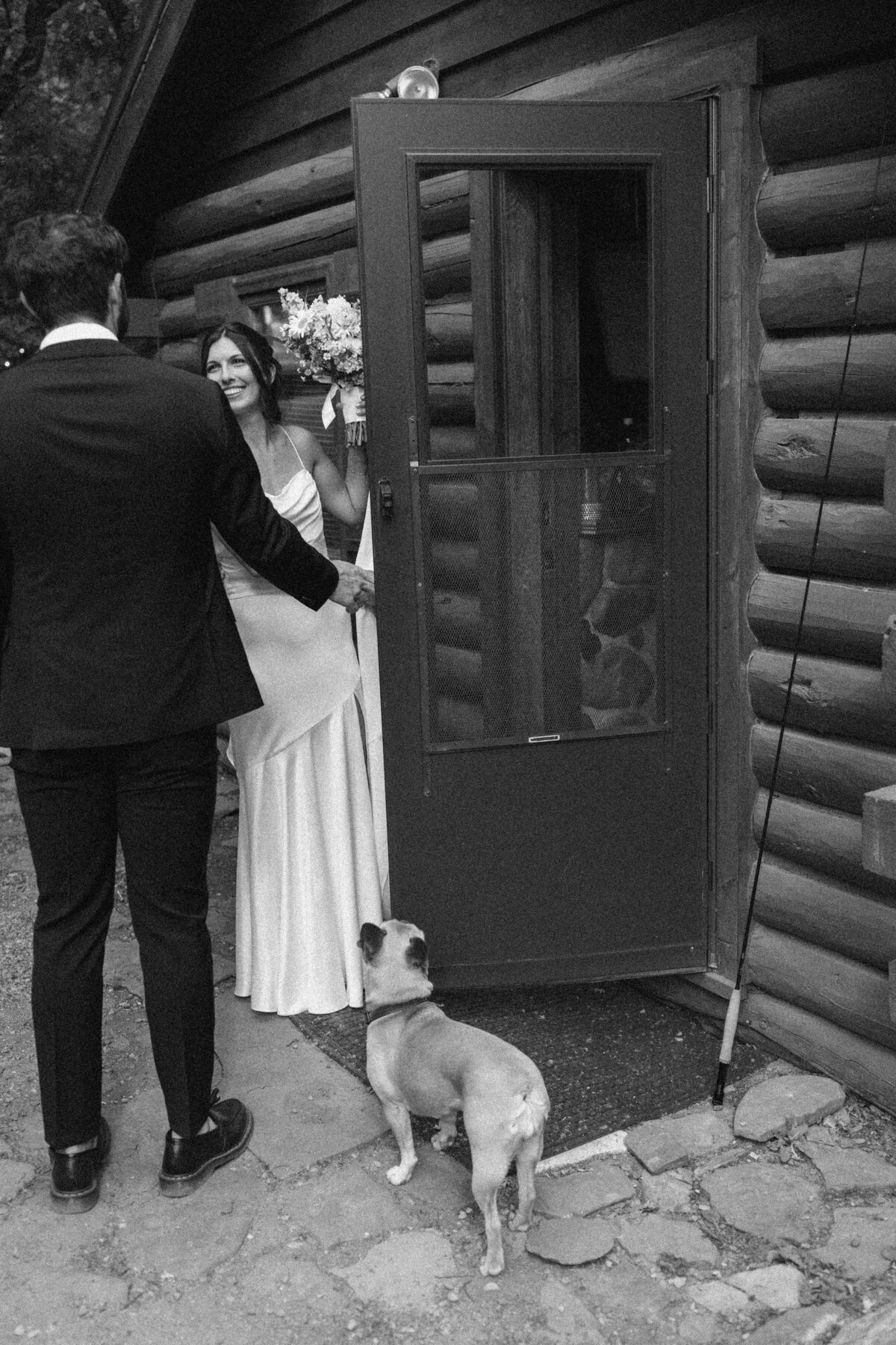 Bride coming out of door looking at groom with small dog looking on