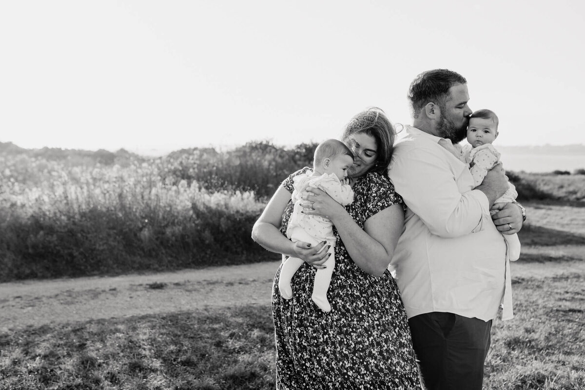 A black and white image of a young mother and father holding their twin daughters at sunset during their photoshoot with Allison Wolf, Boston family photographer