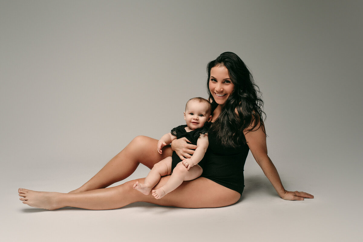 Maternity portrait with mom and toddler. Mom and baby are wearing black body suits while sitting on floor and toddler is on mom's lap