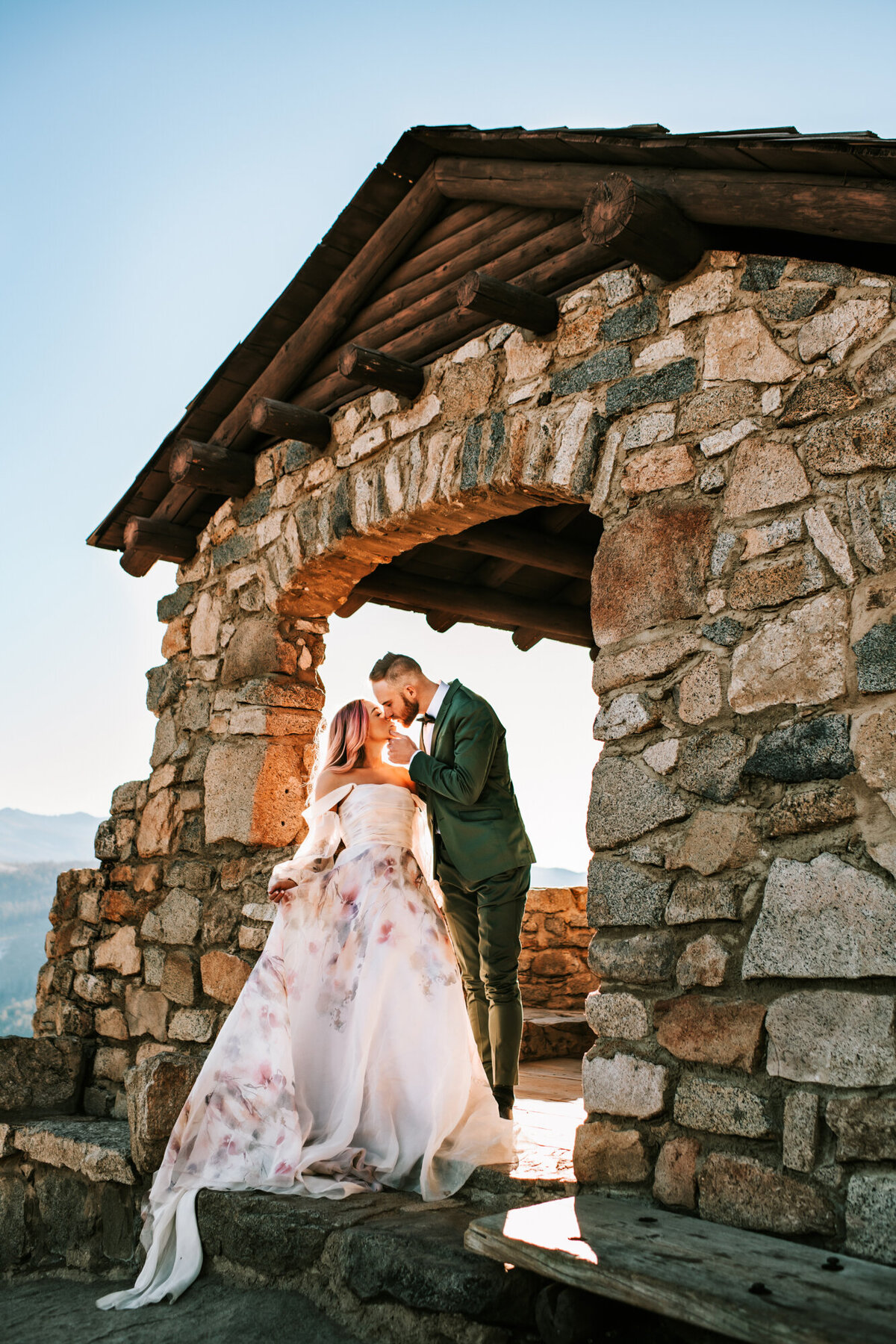 Couples Photography, Man in a blue shirt holding the face of a woman in a wedding dress while they stand in a beautiful stone structure on top of a mountain