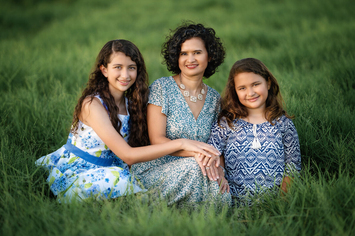 A mom and her daughters and sitting on the levee in Algiers Point Louisiana.  The woman is in the middle.  She is hispanic and has short brown curly hair.  The daughters also have long brown hair.  They are holding hands and are all wearing blue.