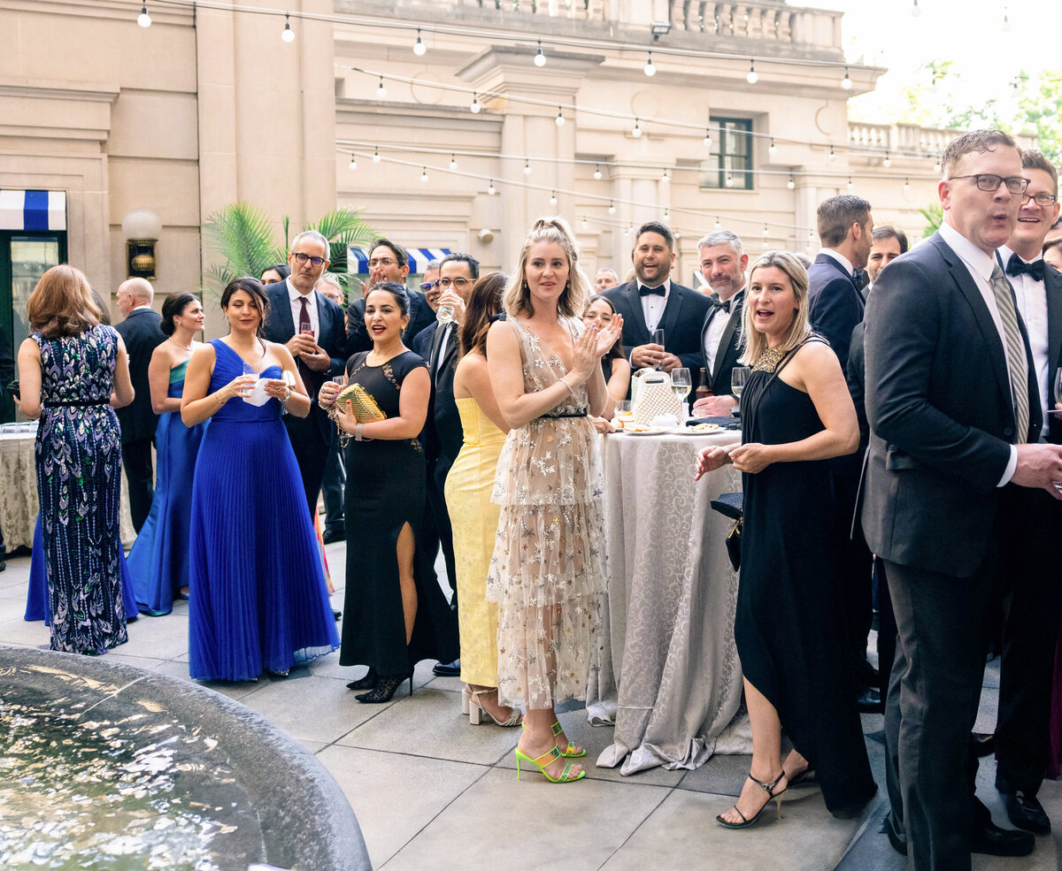 A group of elegantly dressed people gather at an outdoor event near a fountain. Women wear evening gowns, and men are in suits. String lights are overhead, and attendees are socializing around tables with drinks in hand.