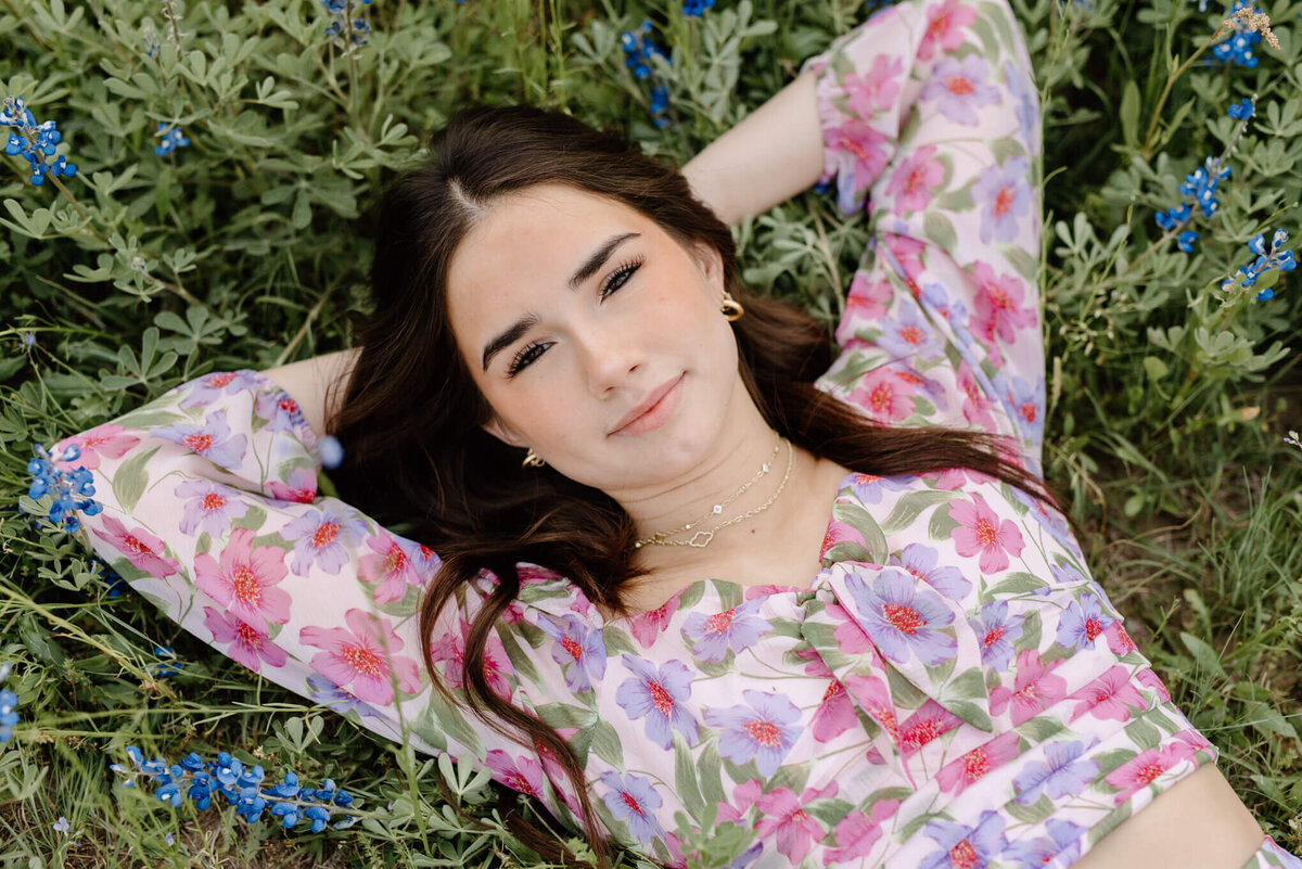 portrait of graduate girl in pink floral dress lying in field of bluebonnets