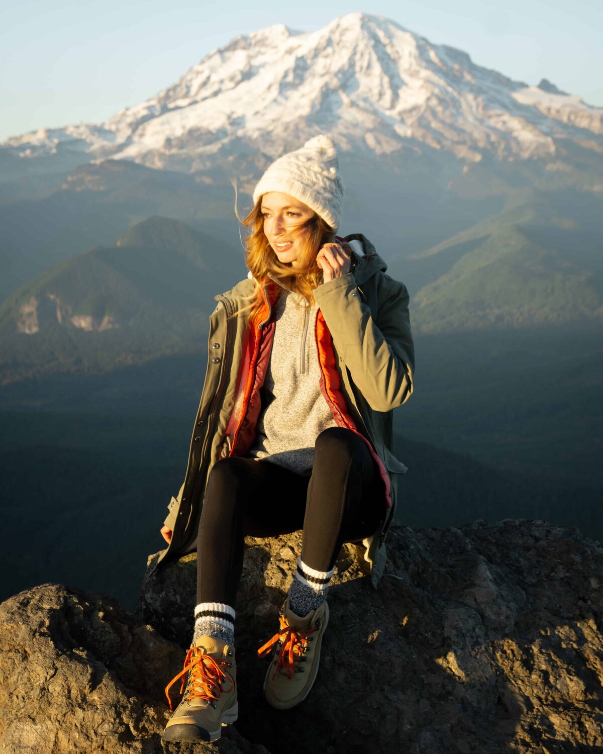 Woman in hiking boots and a hat sitting on a rock with mountains in the background