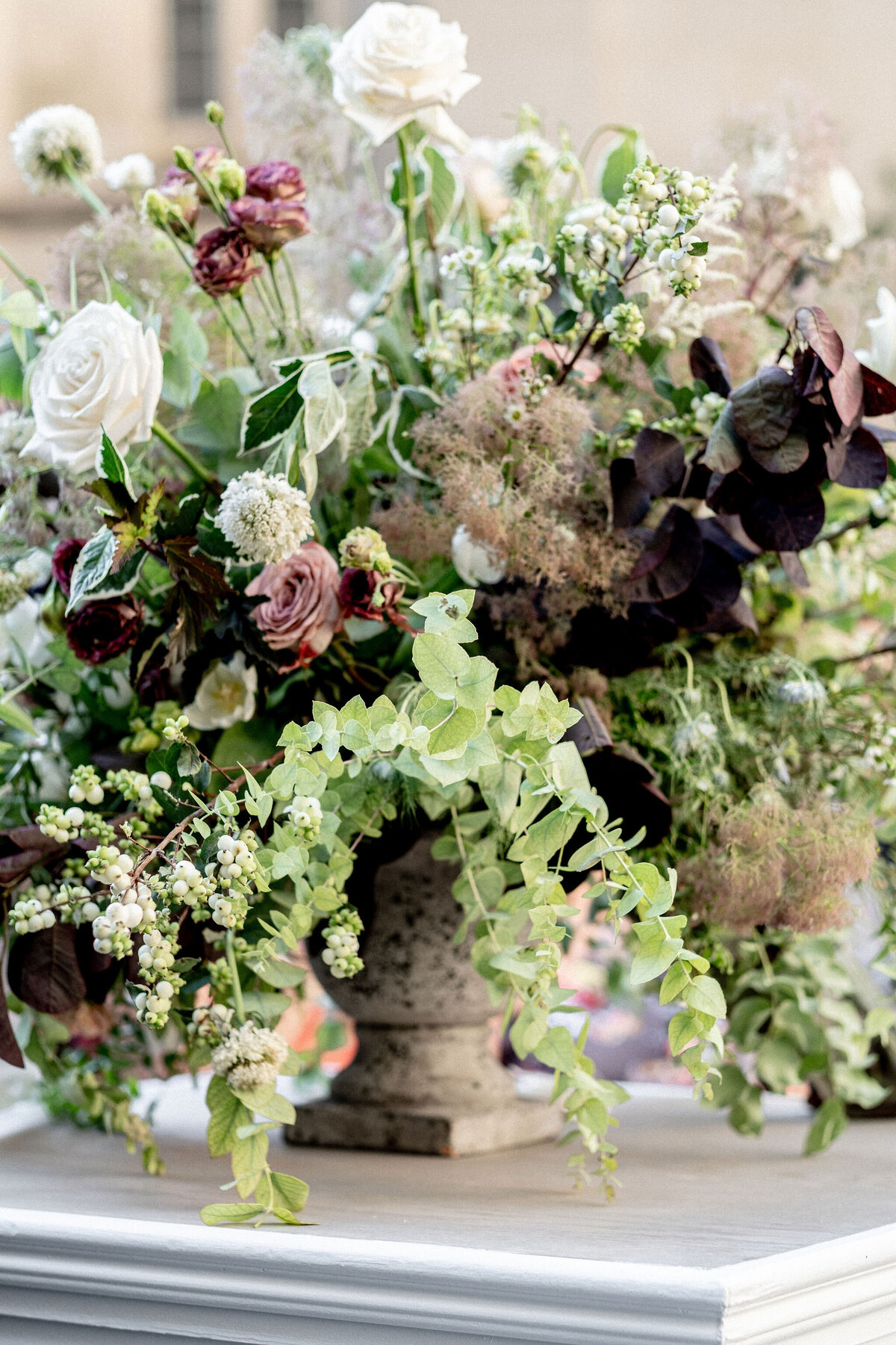 The photo was taken at Washington D.C., Washington National Cathedral by photographer Twah Dougherty. The image captures a decorative stone vase with an arrangement of various flowers and greenery. The bouquet includes white roses, purple flowers, and assorted foliage, creating a lush and vibrant display on a white surface. Current Films is a Videographers company that could capture the beauty of this arrangement through video.