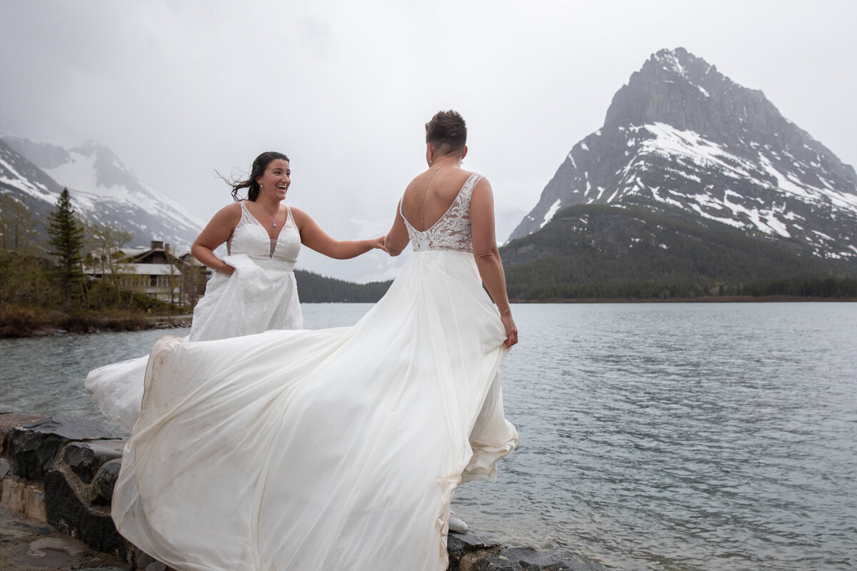 Two brides in flowing white dresses laugh and twirl next to a glacial lake surrounded by towering mountains and 
