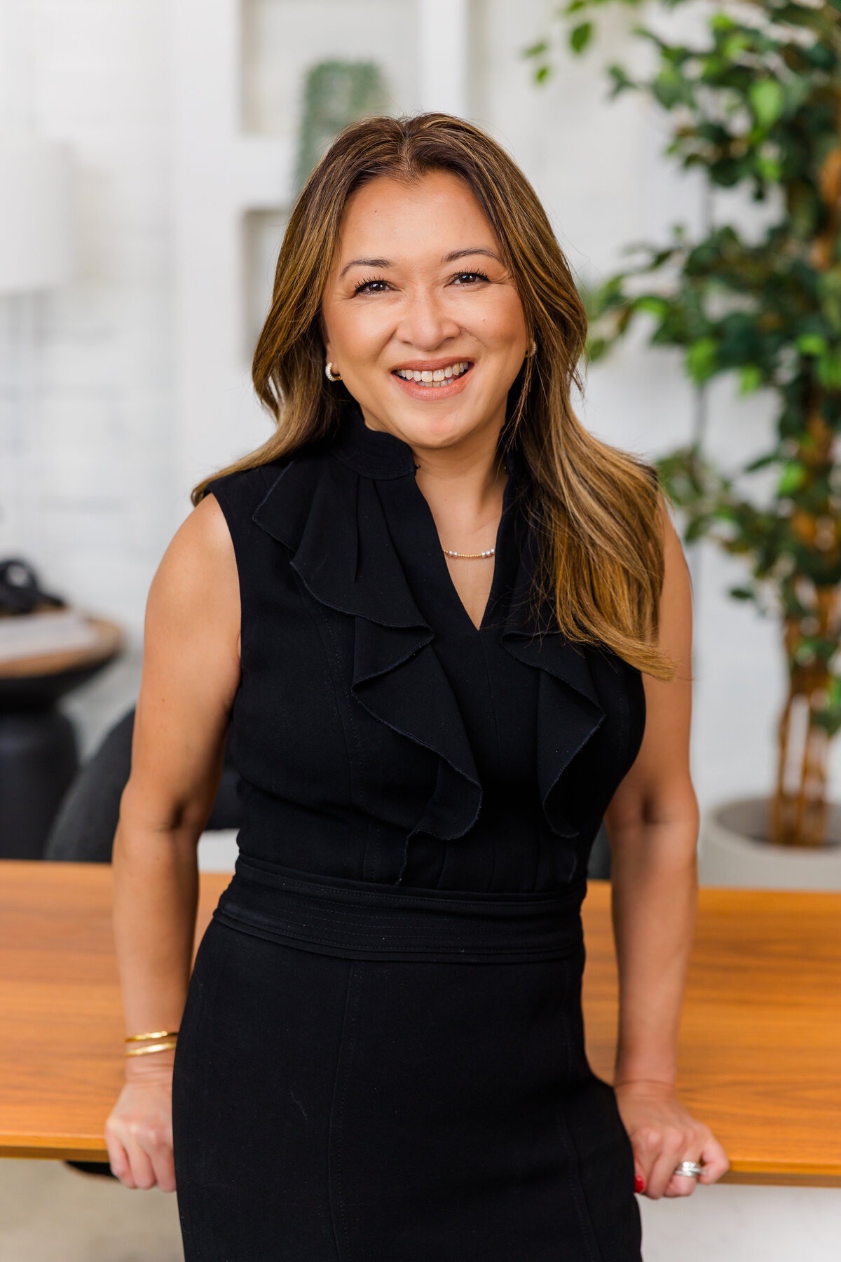 Corporate portrait of woman wearing a black dress leaning against an office desk with a lifestyle background