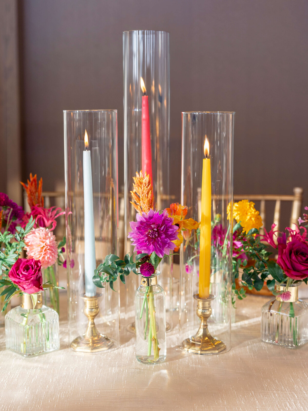 A decorative arrangement featuring tall candles in glass holders and vibrant flowers in small vases. The candles are red, blue, and yellow, surrounded by roses and other colorful blooms, creating an elegant, festive display on a table.