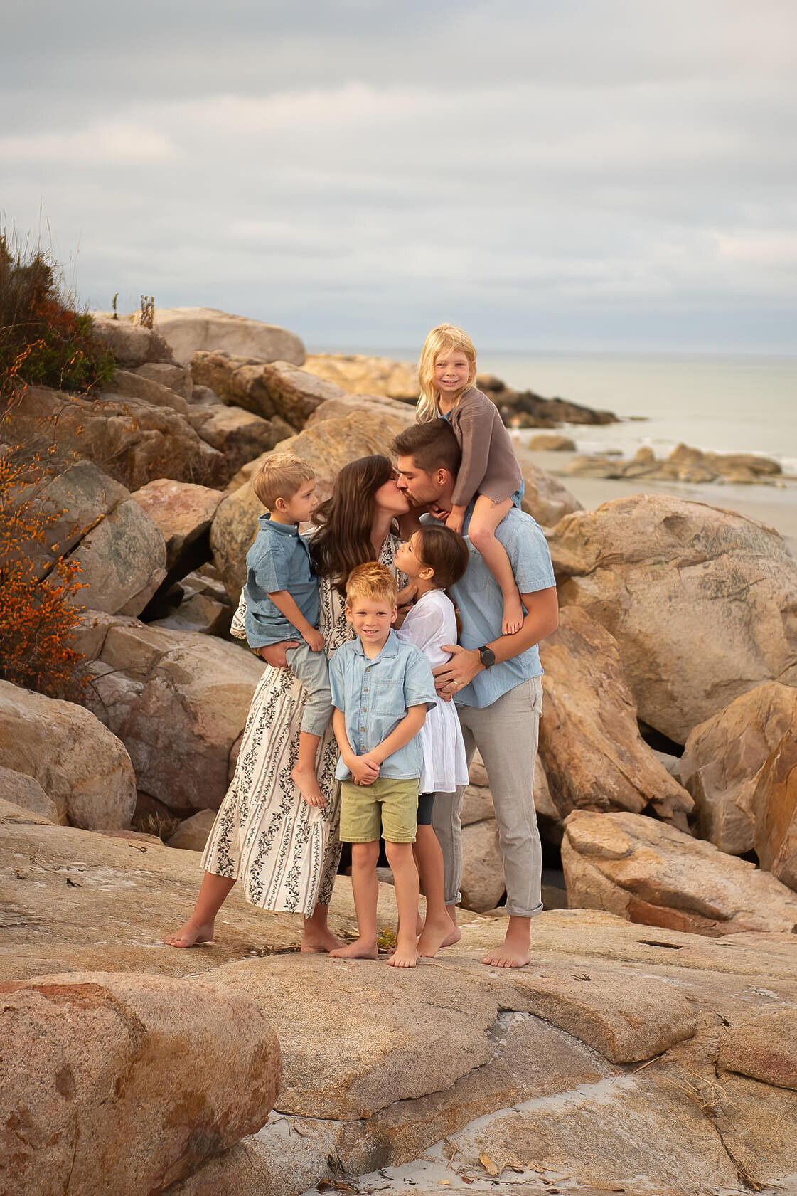 A family of six stand on a rock at Wingaersheek beach with the mom and dad kissing while one little girl sits on her dads shoulders and the other kids look on.