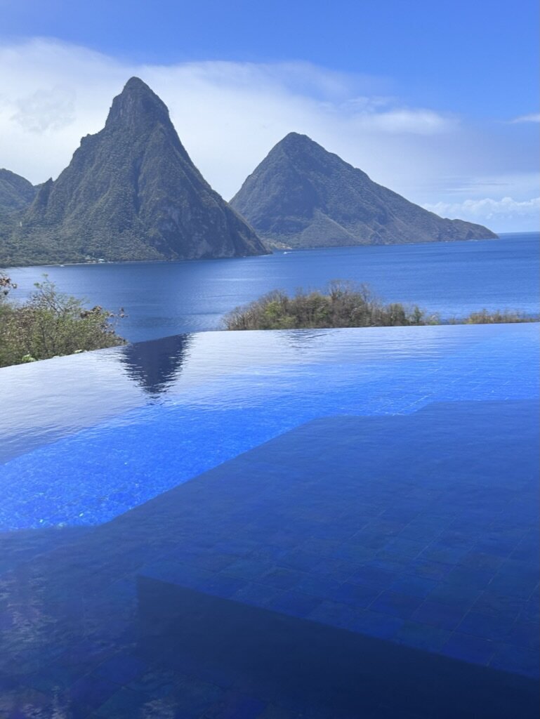 An infinity pool overlooking the ocean with two tall, mountainous peaks in the background. The scene is set under a clear blue sky, showcasing a tranquil and picturesque destination.