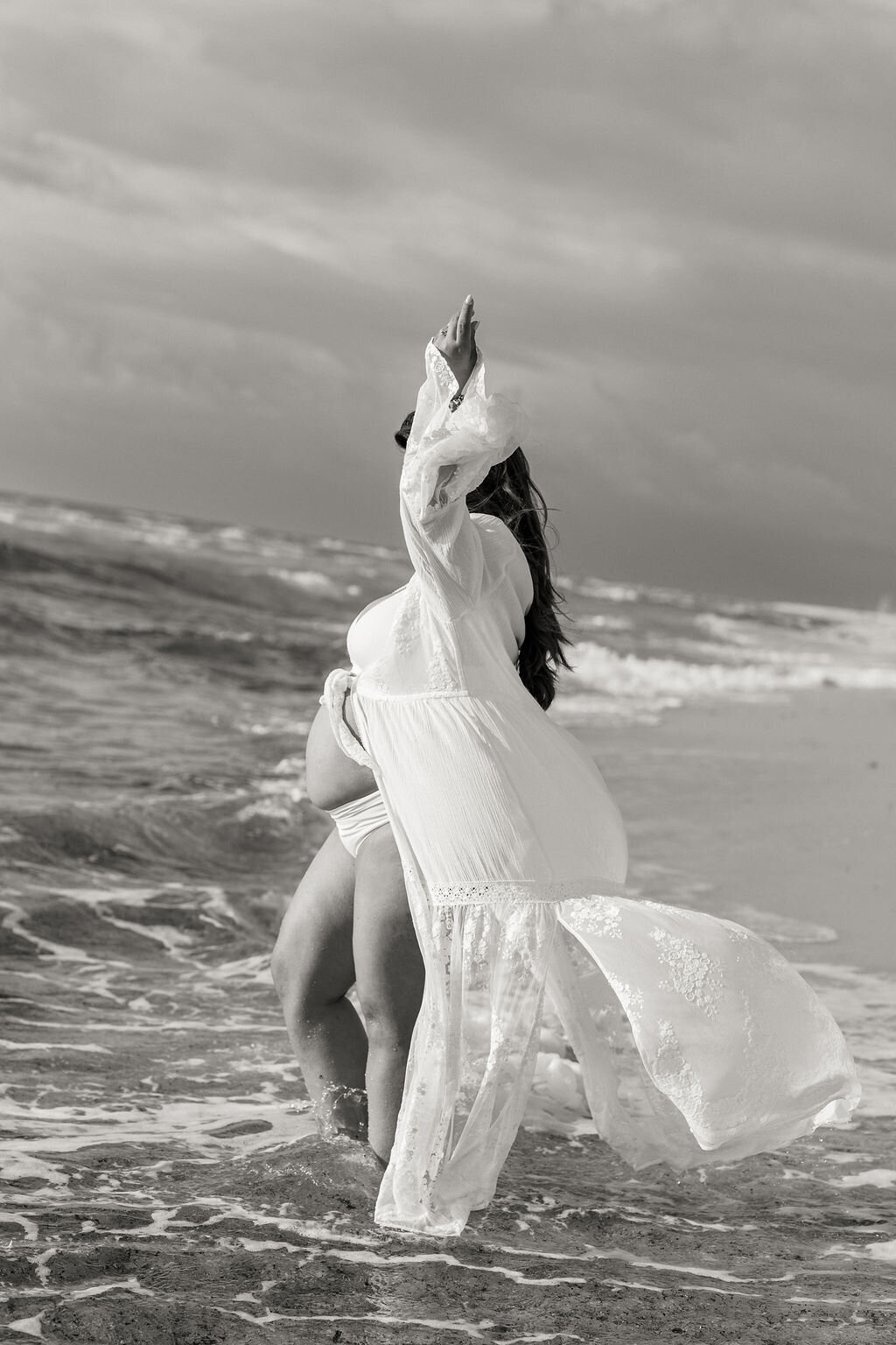 Pregnant woman in lace robe stepping into ocean waves at Miami beach, raising arm in serene pose, black and white image.