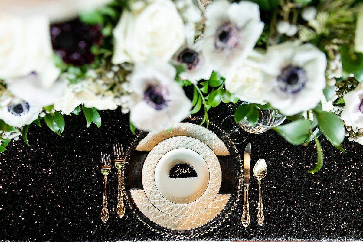 Sitting on a black glittered table cloth is a clear glass charger stacked with quilted ivory plates wrapped with a black satin napkin and topped with a black agate  slice seating card. On either side of the plate is silverware and at the top are a large black, white and green floral centerpiece of anemone at Noah Liff Opera Center Wedding.