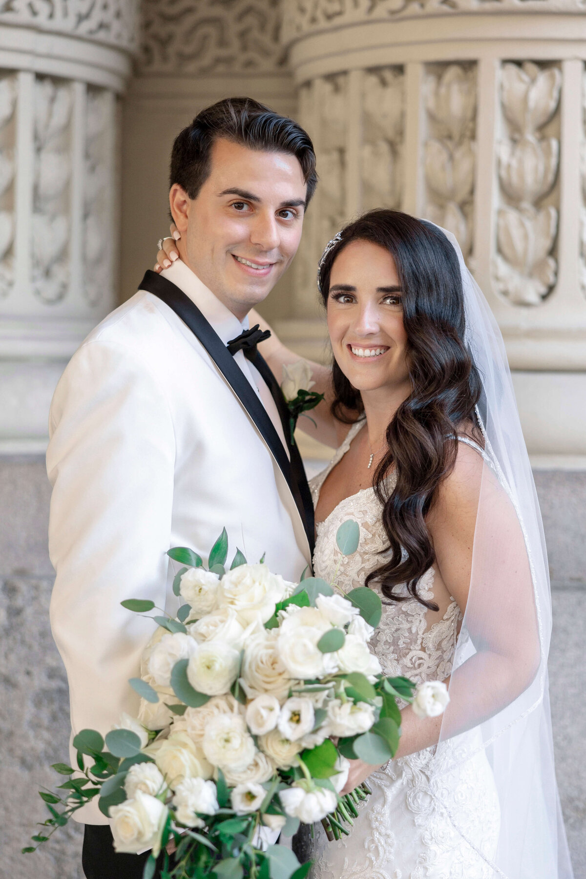 A bride and groom stand closely together, smiling. The groom wears a white tuxedo with a black bow tie, while the bride wears a lace wedding dress and veil, holding a bouquet of white flowers. They are set against an ornate stone background.