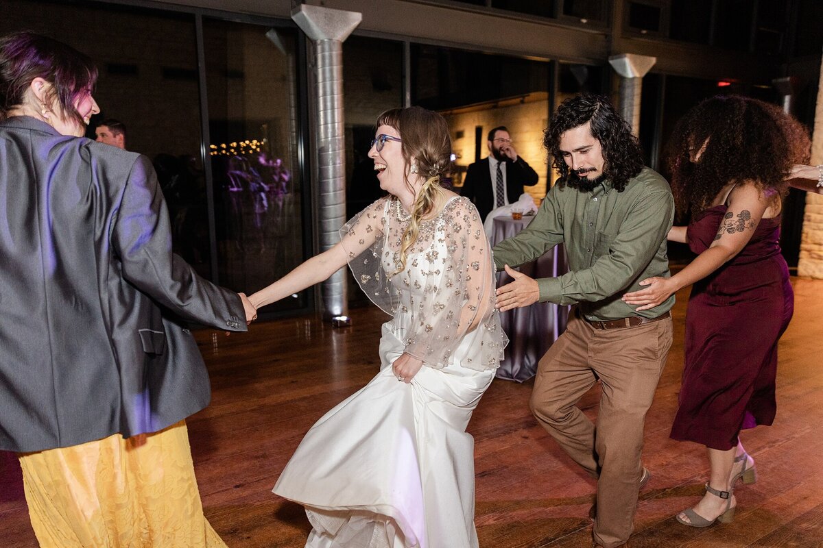 Photo of a bride taking part in a conga line during her wedding reception at the Lady Bird Johnson Wildflower Center in Austin, Texas. The bride is in the center of the photo and is wearing an elegant, long, white dress. Her guests continue the line before and after her and lead her along in celebration.