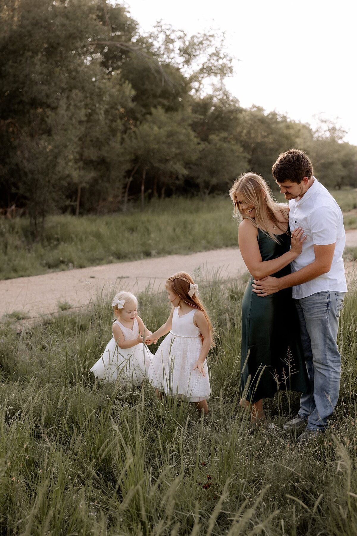 mom and dad standing in a field with arms wrapped around each other looking down at their two daughters playing.