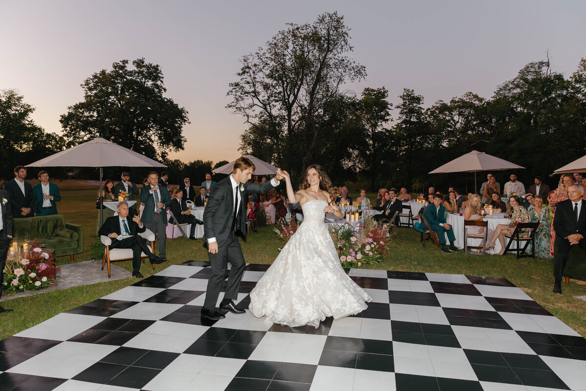 Couple sharing their first dance on a black-and-white checkered dance floor during an outdoor wedding reception, surrounded by candlelit tables and lush floral arrangements.