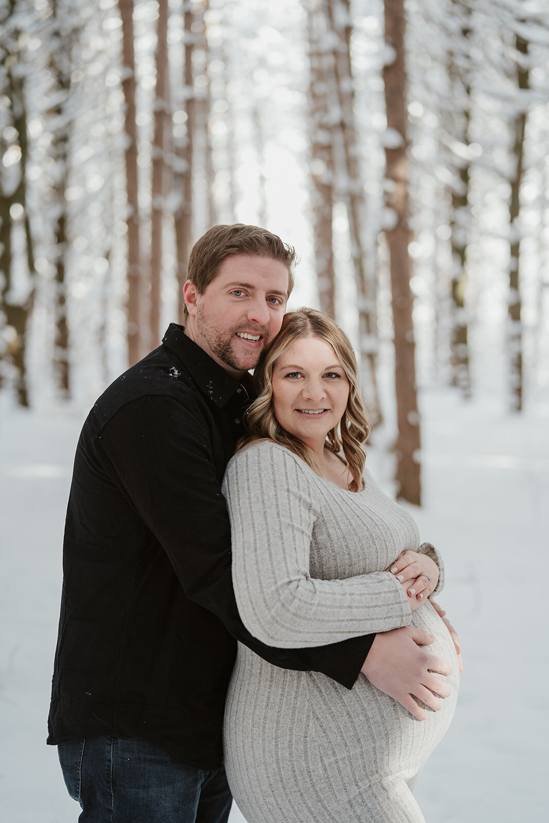 Pregnant woman posing in front of snow-covered pine trees at Al Sabo Preserve during her winter maternity session in Kalamazoo.