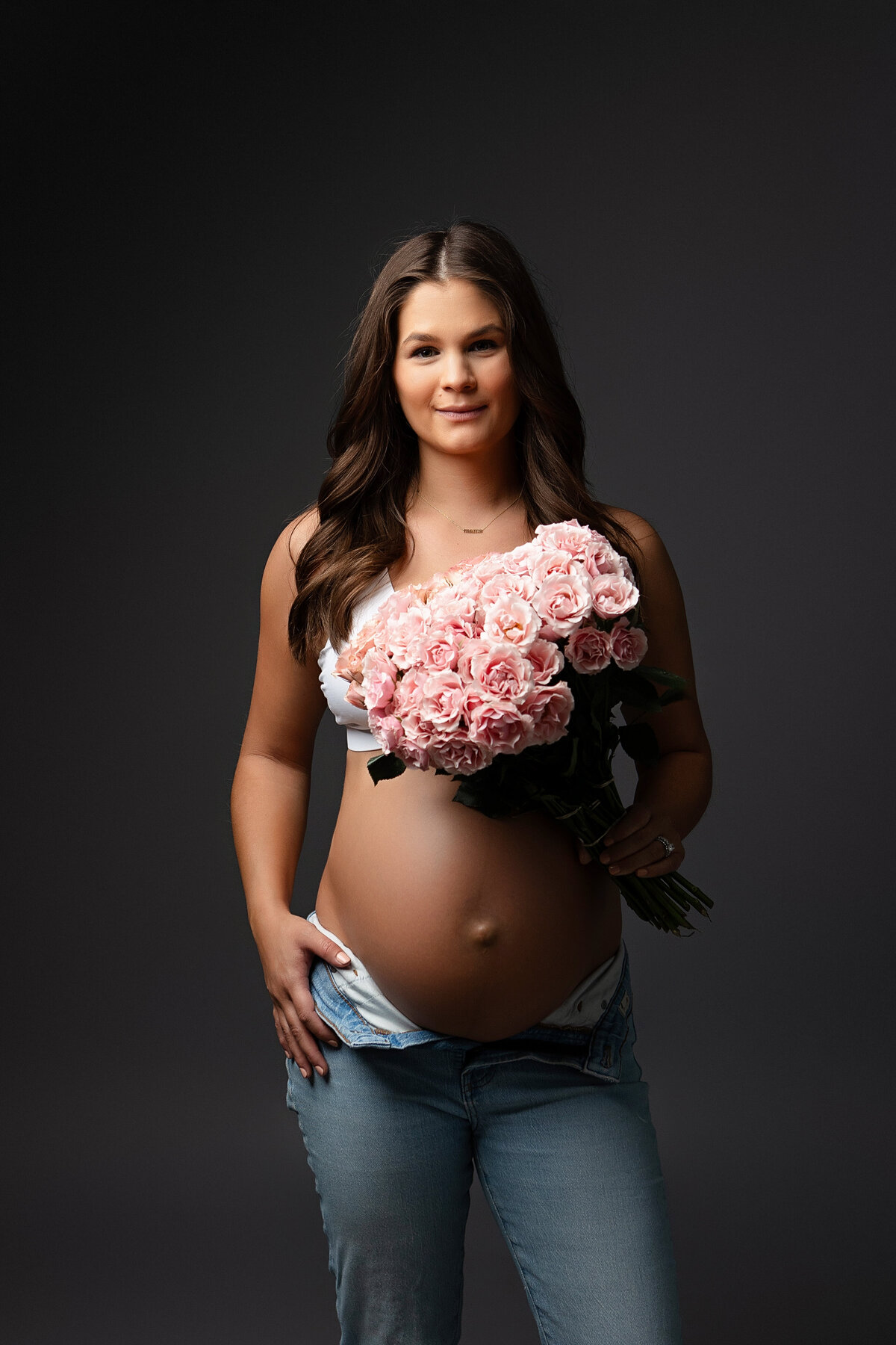 Expectant mother holding pink flowers over pregnant belly while wearing jeans during a Fresno, CA photoshoot