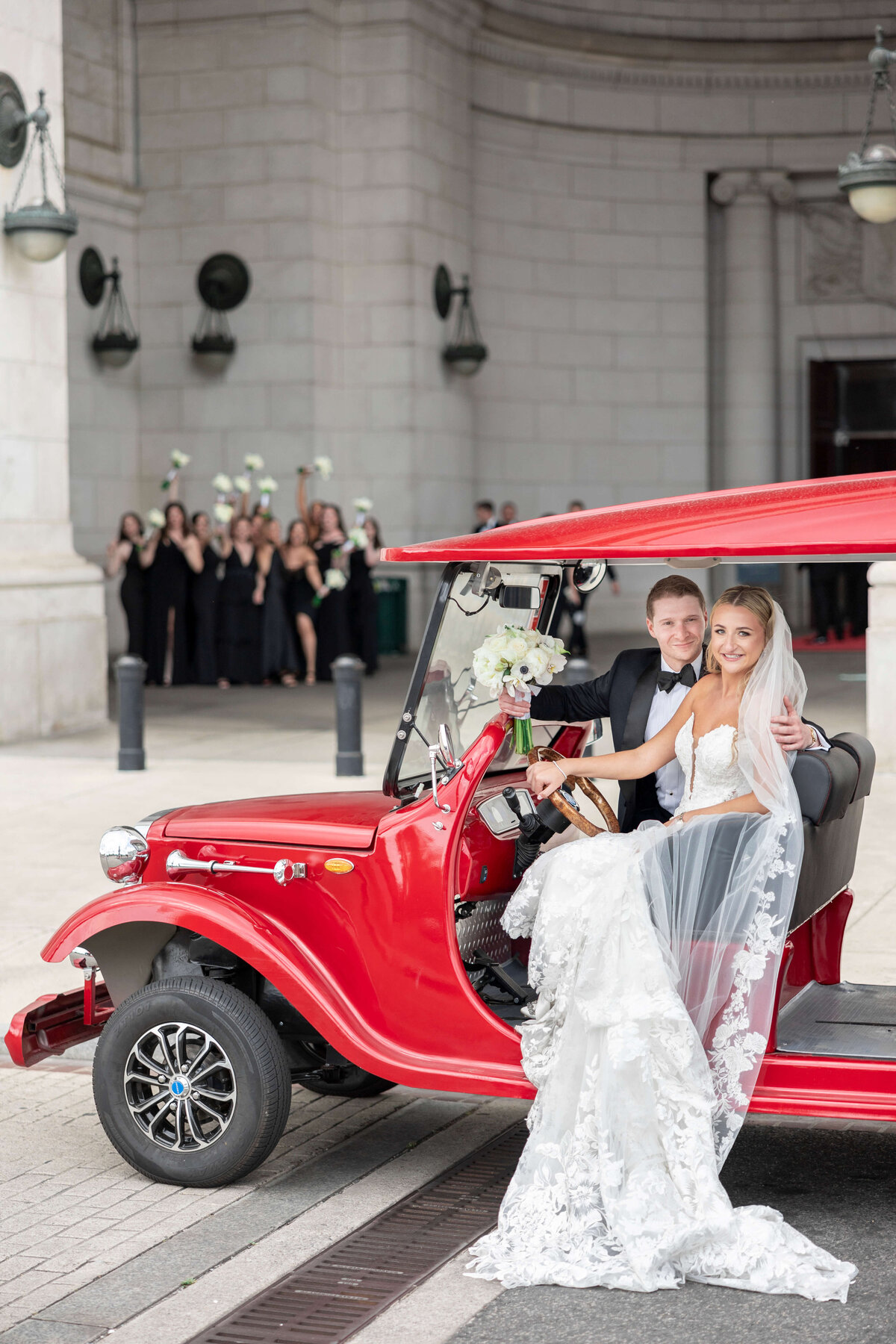 A bride and groom, smiling, sit in a vintage red car outside a grand building. The bride holds a bouquet, and the groom is in a black tuxedo. In the background, a group of people dressed in black celebrate, raising white flowers.