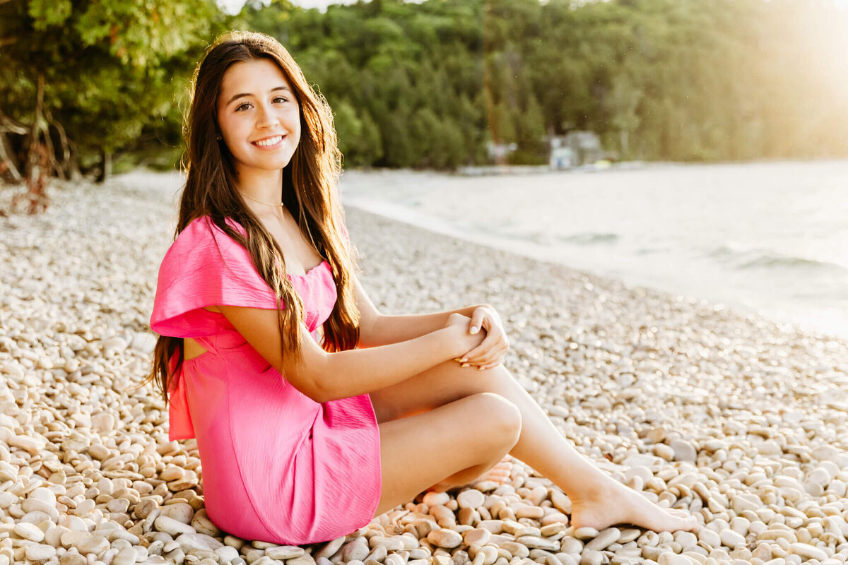 beautiful teen girl in a bright pink dress resting hands on her knees and smiling for her Appleton senior photo session