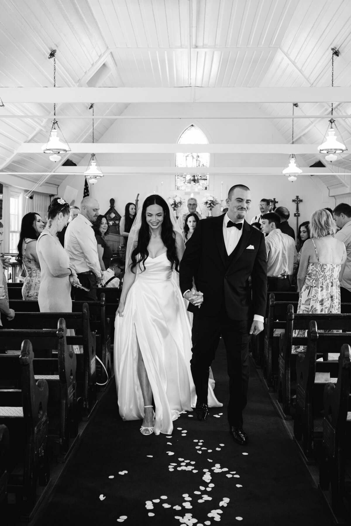 excited bride and groom exiting white chapel after ceremony with cheering guests in black and white